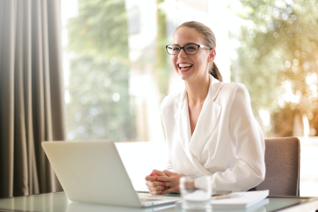 risingqueen- Lächelnde Geschäftsfrau, die in einem hell erleuchteten Büro an einem Laptop an ihrer Marketingstrategie arbeitet.